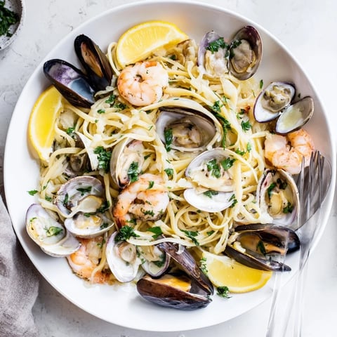 A close-up of Seafood Linguine in a white bowl, featuring plump shrimp, open clams, and mussels glistening in a white wine garlic sauce, with fresh parsley and lemon wedges.