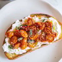A close-up of Tomato Ricotta Toast with golden sourdough, creamy ricotta, and glistening roasted cherry tomatoes, garnished with fresh basil and flaky sea salt.