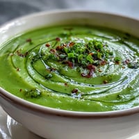 A bowl of vibrant Courgette, Pea and Pesto Soup garnished with fresh basil, served beside a slice of crusty bread for dipping.
